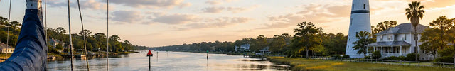 Golden hour sailing scene along the Atlantic Intracoastal Waterway featuring a calm coastal channel, lighthouse, marsh shoreline, waterfront homes, and a sailboat traveling through protected eastern seaboard cruising waters.