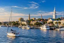 Charleston Harbor marina with sailboats, historic waterfront homes, palm trees, and calm coastal water beneath a warm southern sunset sky.