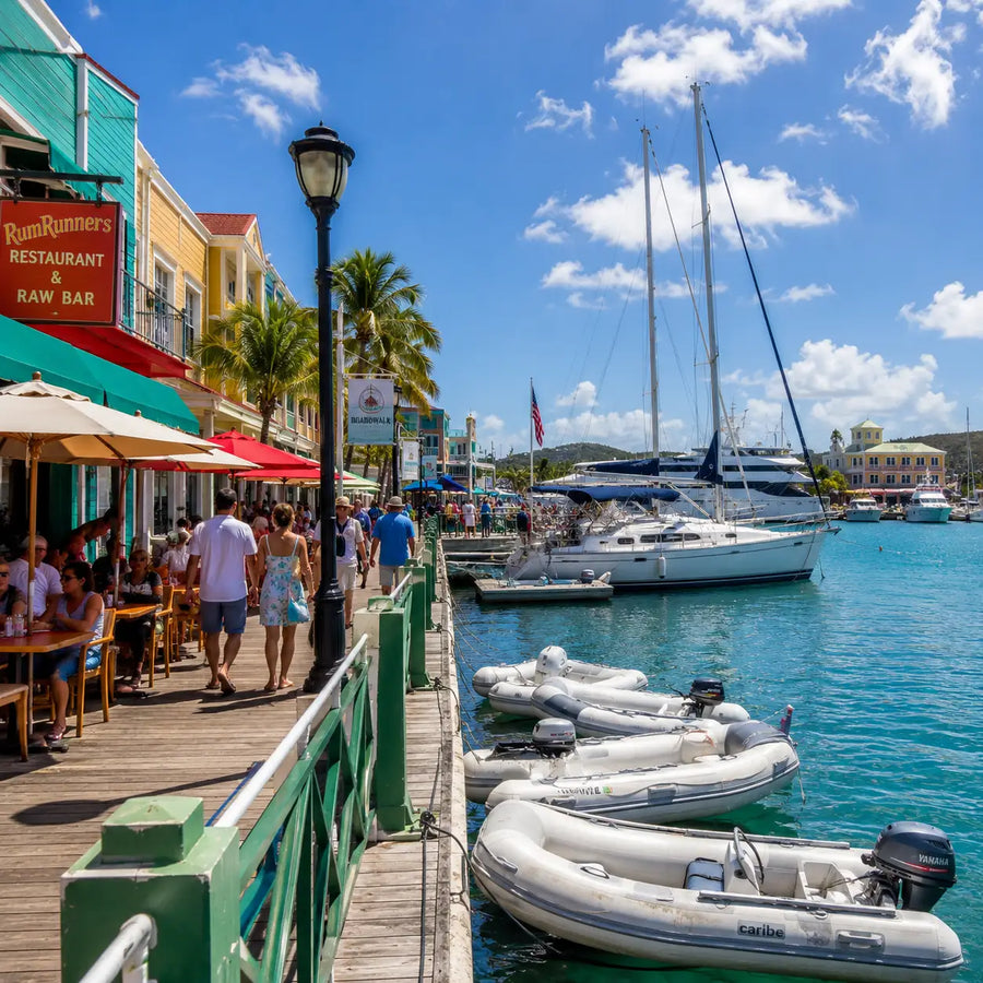 The lively Christiansted waterfront boardwalk on St. Croix featuring restaurants, shops, dinghies, visiting yachts, palm trees, and pedestrians enjoying the historic harbor atmosphere.