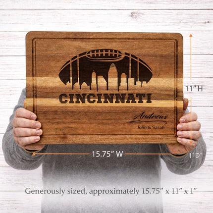 Cincinnati football cutting board shown in hands for size reference, measures 15.75 x 11 x 1 inches, solid acacia wood with laser engraved skyline and custom name