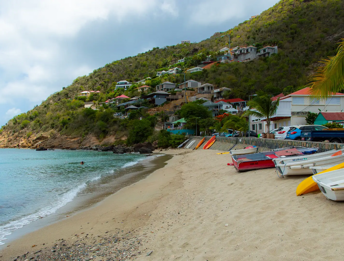 Traditional fishing boats resting along the beach at Corossol Harbor in St. Barths, with waterfront homes and hillside village scenery overlooking the Caribbean Sea.