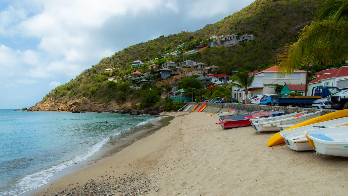 Traditional fishing boats resting along the beach at Corossol Harbor in St. Barths, with waterfront homes and hillside village scenery overlooking the Caribbean Sea.
