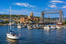 Duluth Harbor on Lake Superior with sailboats, historic canal waterfront, lighthouse pier, and rugged northern shoreline beneath a bright summer sky.