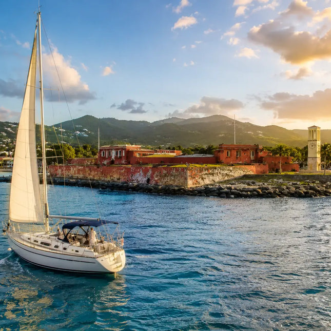 Sailboat departing Frederiksted Harbor along the west coast of St. Croix, U.S. Virgin Islands, with Fort Frederik, turquoise Caribbean waters, and lush green hills in the background.
