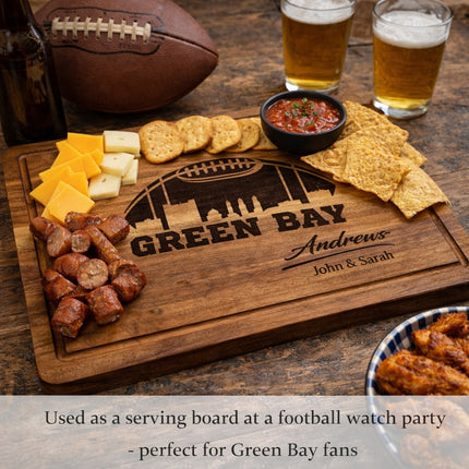 Personalized Green Bay football cutting board used as a serving board with snacks during a game day gathering, showing dark laser engraved skyline and family name
