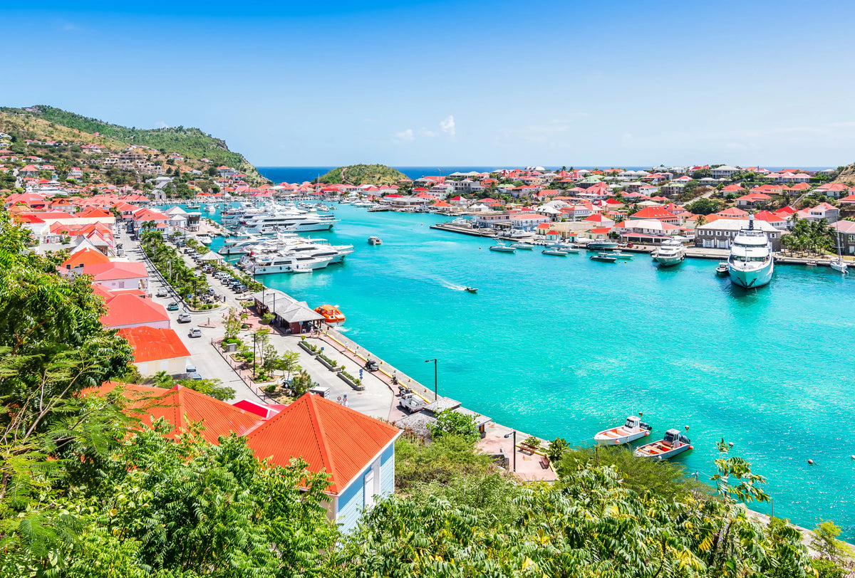 Luxury yachts and turquoise waters in Gustavia Harbor, St. Barths, featuring the waterfront marina and red-roofed Caribbean buildings.