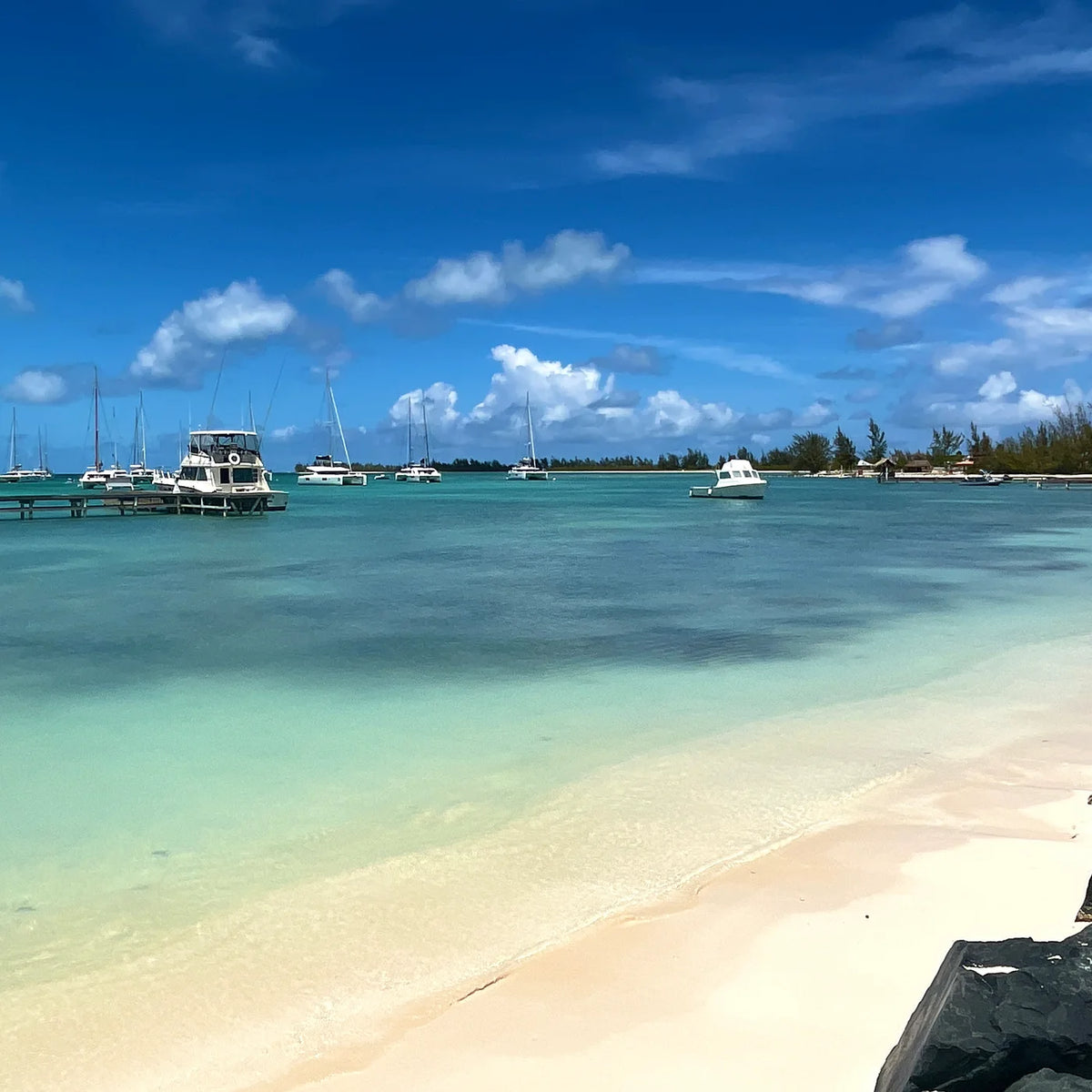 Sailboats and powerboats offshore from a dock, with pale sand and shallow turquoise water in the foreground.