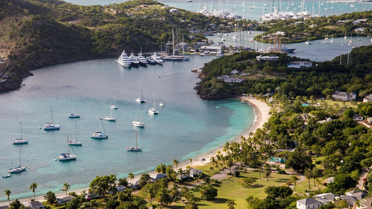 Elevated view of English Harbour, Antigua, with sailboats, waterfront docks, a curved beach and wooded hills.