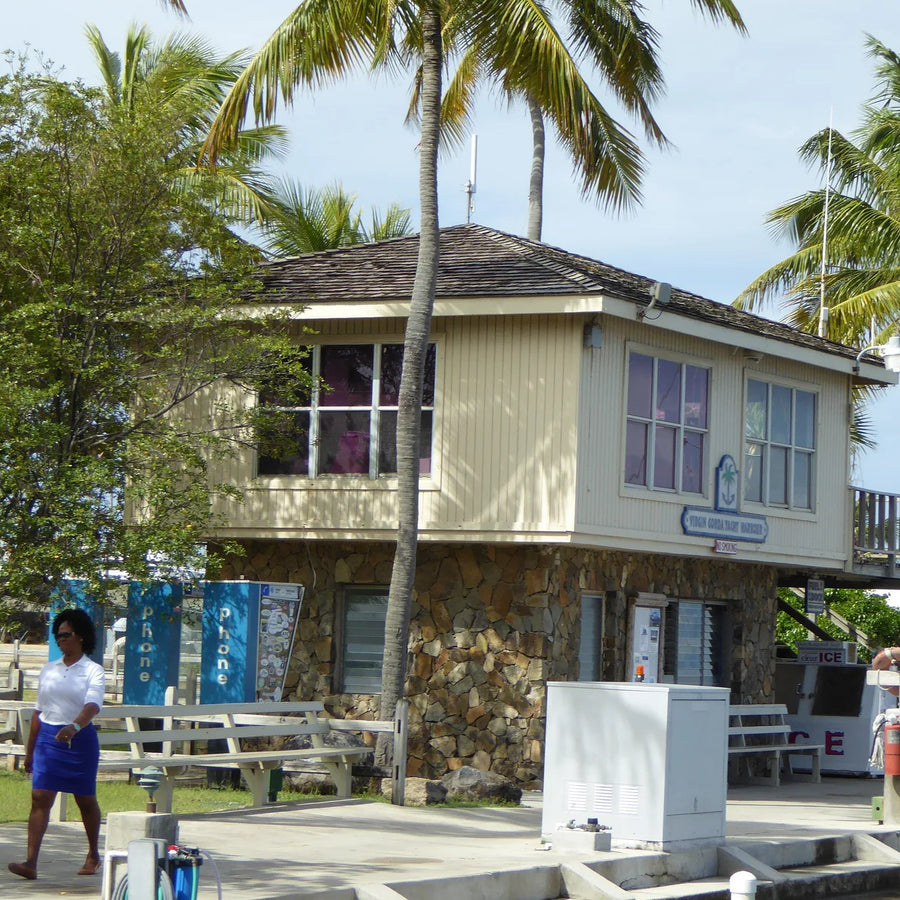 A cream-colored waterfront building with a stone lower level, palm trees, benches and a yacht alongside the dock.