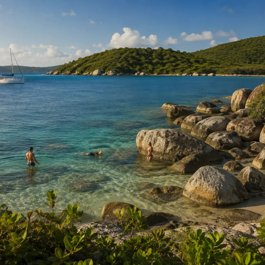People swim beside rounded granite shoreline boulders in clear blue water, with a sailboat and green hills beyond.