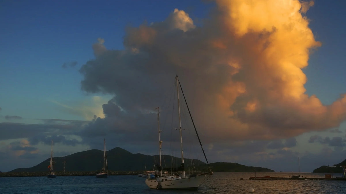 A sailboat on dark blue water beneath towering gold-lit clouds, with low green hills in the background.