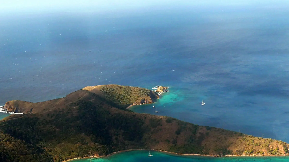 Hilly coastal headlands bordered by turquoise shallows and deeper blue water, with several sailboats offshore.