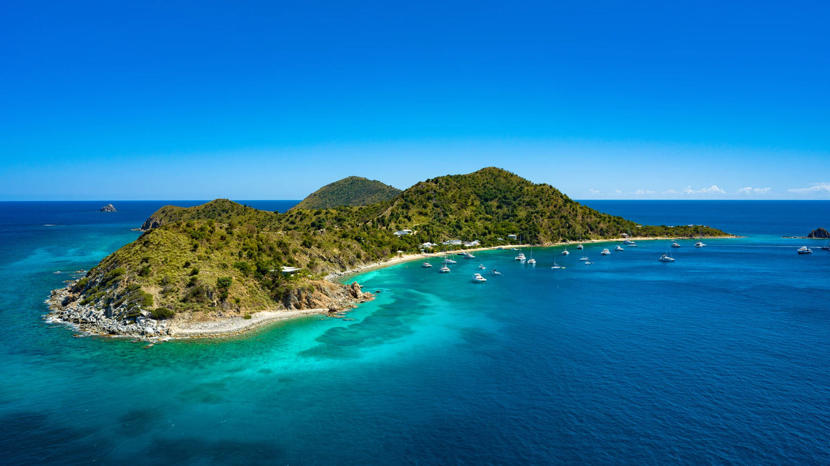 Aerial view of a green, hilly island with a narrow beach, rocky foreground headland and sailboats and powerboats across blue-green water.