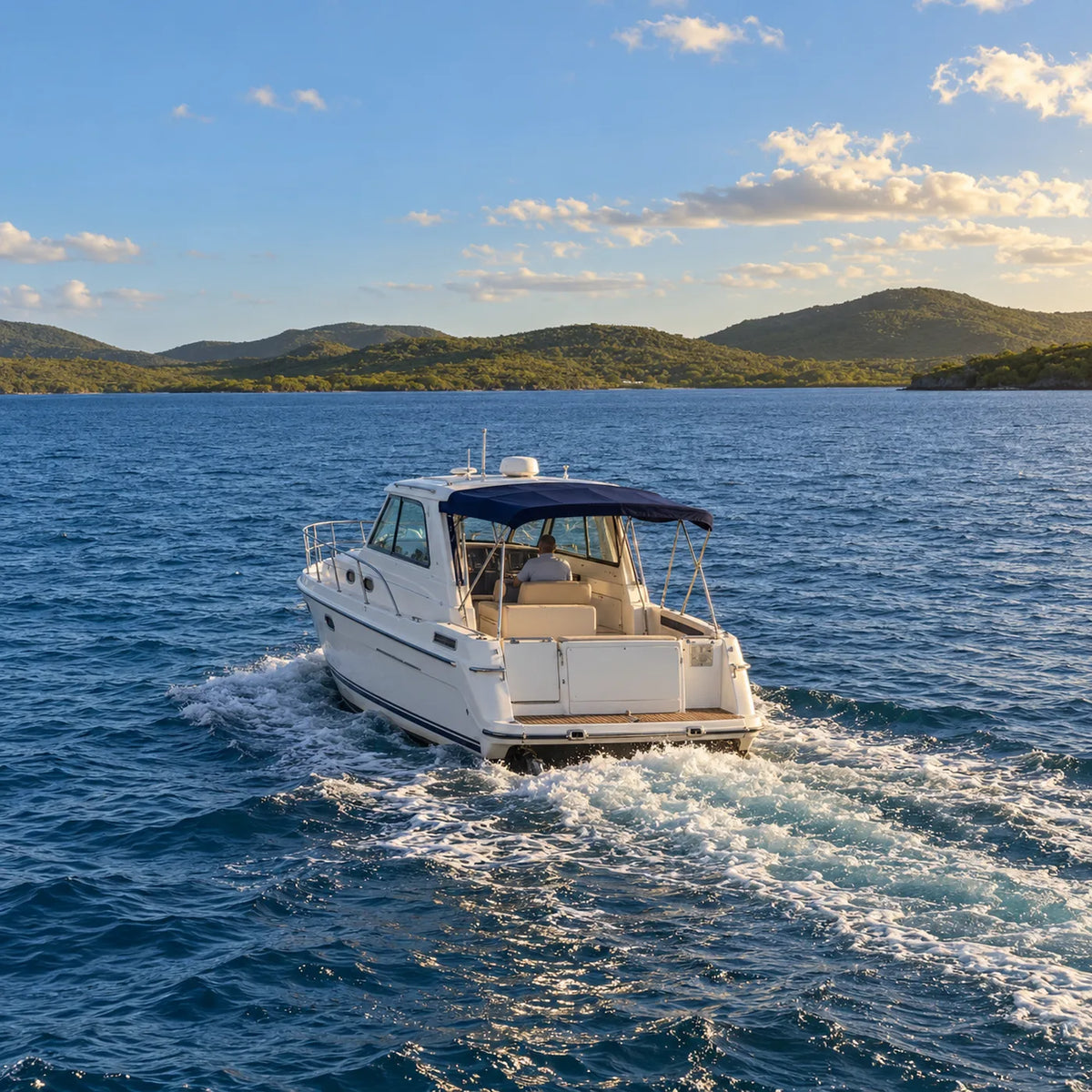Stern view of a white cabin cruiser with a navy canopy, leaving a foamy wake across blue water toward green hills.