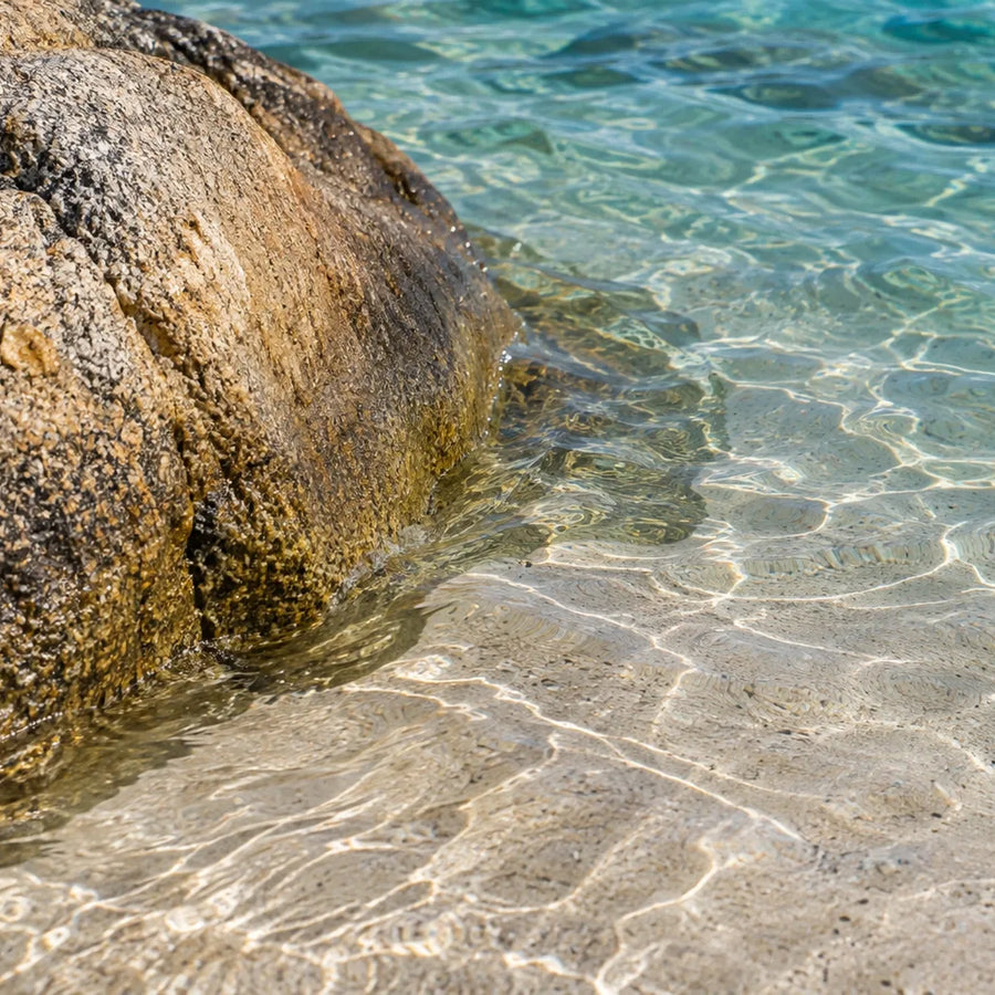 Close view of textured granite beside clear shallow water, with rippled light patterns on the sandy bottom.