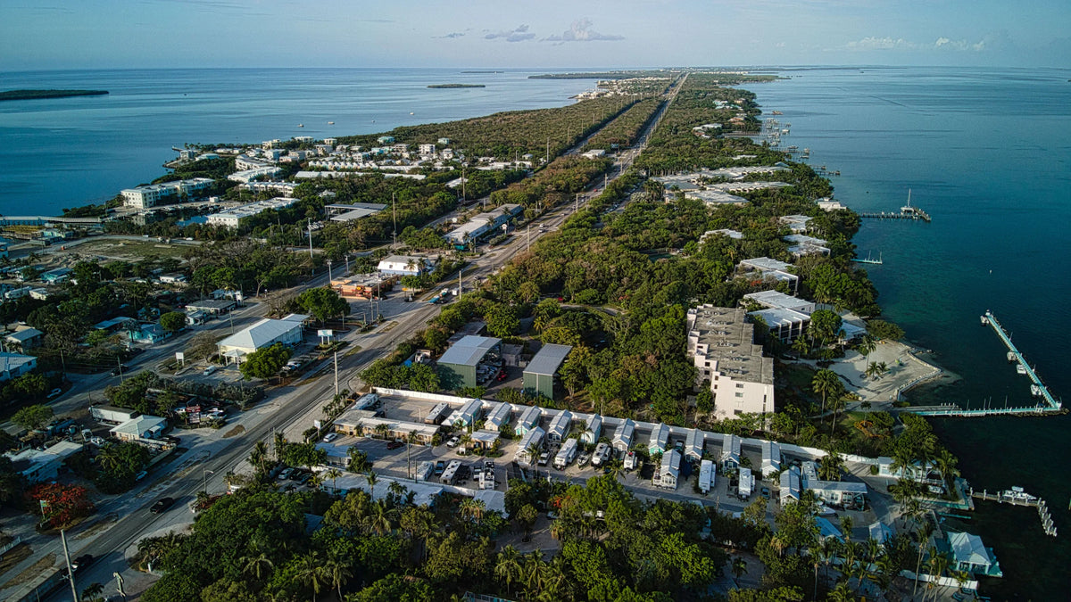 A road runs through a tree-covered island, with waterfront buildings and docks beside blue water on both sides.