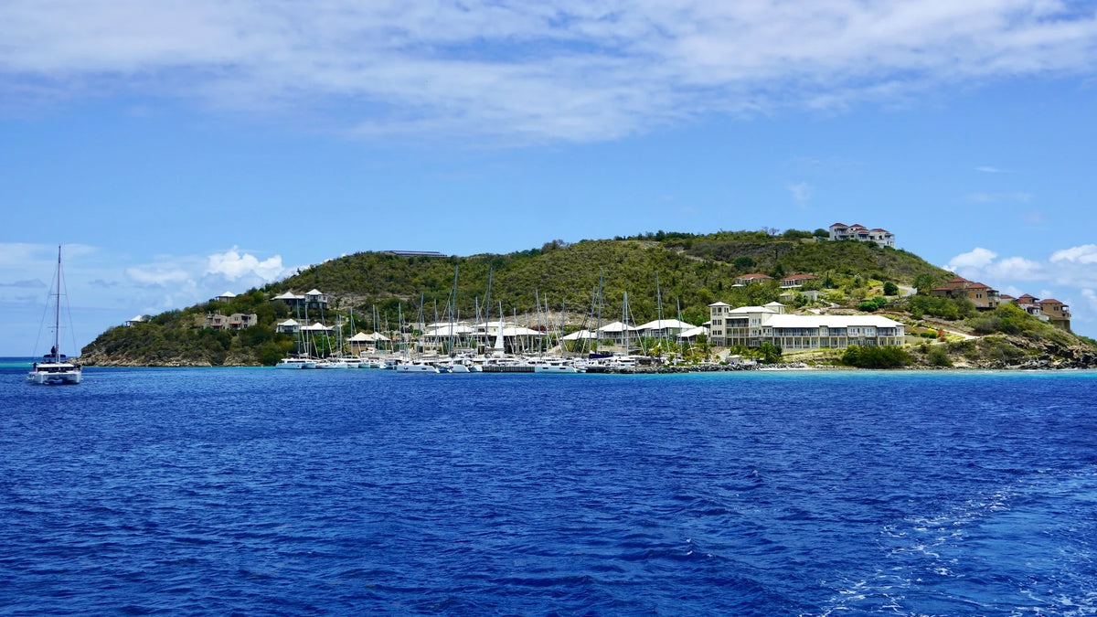 Blue water in the foreground, sailboats and waterfront buildings along a green island hillside, and scattered clouds overhead.