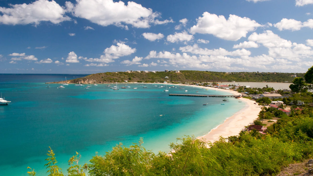 Elevated view of a turquoise bay, scattered boats, a long pier, beachfront buildings, and green coastal slopes.
