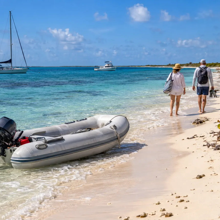 An inflatable dinghy rests at a sandy shoreline as two boaters walk along the beach, with a sailboat and power cruiser offshore.