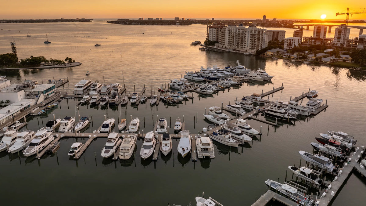 Aerial sunset view of yachts docked in a marina beside Sarasota’s waterfront buildings, with open bay water beyond.