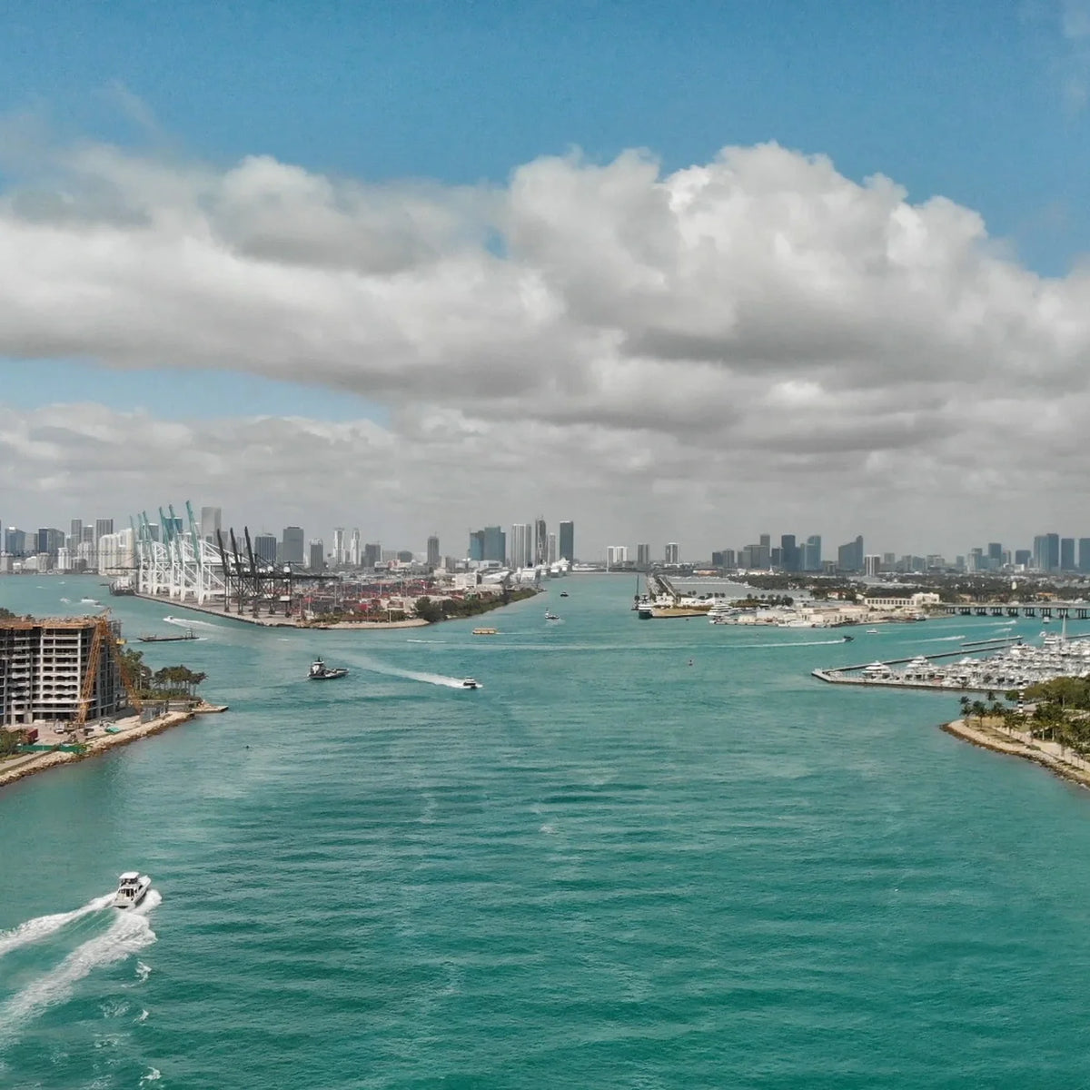 A powerboat leaves a white wake across blue-green water, with marina docks, port cranes and a distant skyline ahead.