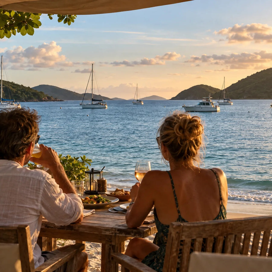 Two people dine beside a sandy shore at golden hour, overlooking anchored sailboats and a power cruiser.