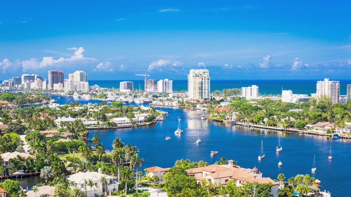 Sailboats sit on blue water between palm-lined homes, with marina docks, coastal high-rises and the ocean beyond.