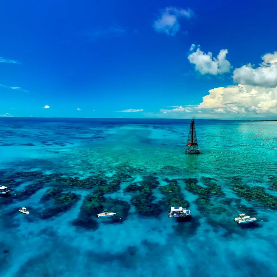 Elevated view of a skeletal lighthouse and several boats above dark reef patches in turquoise water beneath large white clouds.