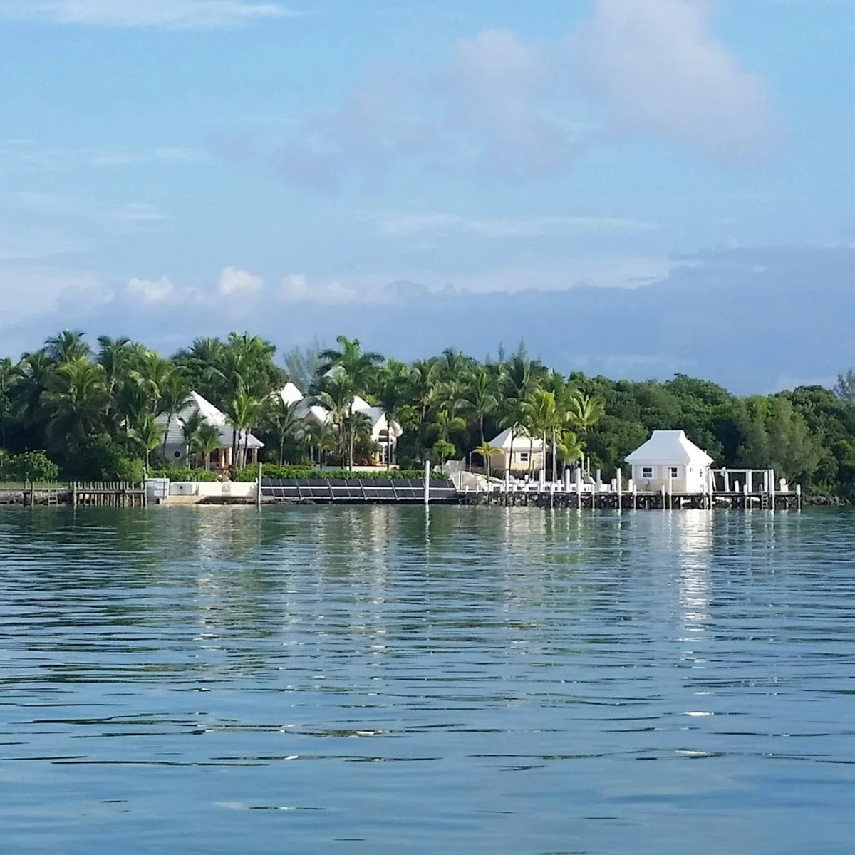 White-roofed waterfront homes among palms, with a small dock building and rippled water in the foreground.