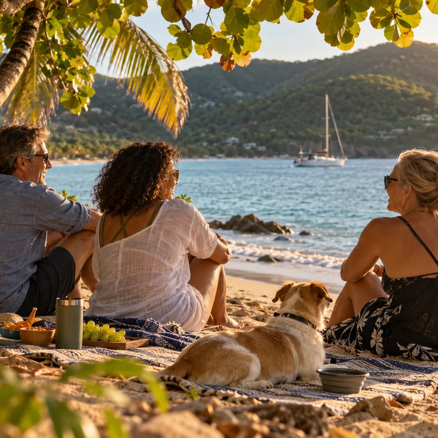 Three adults and a small dog sit beneath coastal trees beside the water, with cruising boats and green hills in the distance.