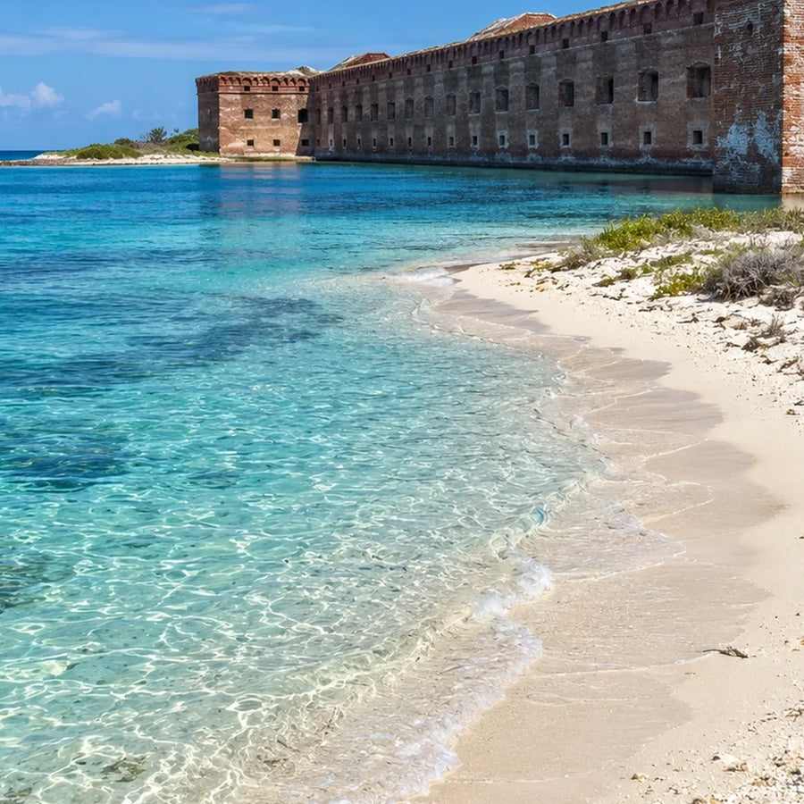 Clear turquoise water laps a pale sandy beach beside a large weathered brick fort.