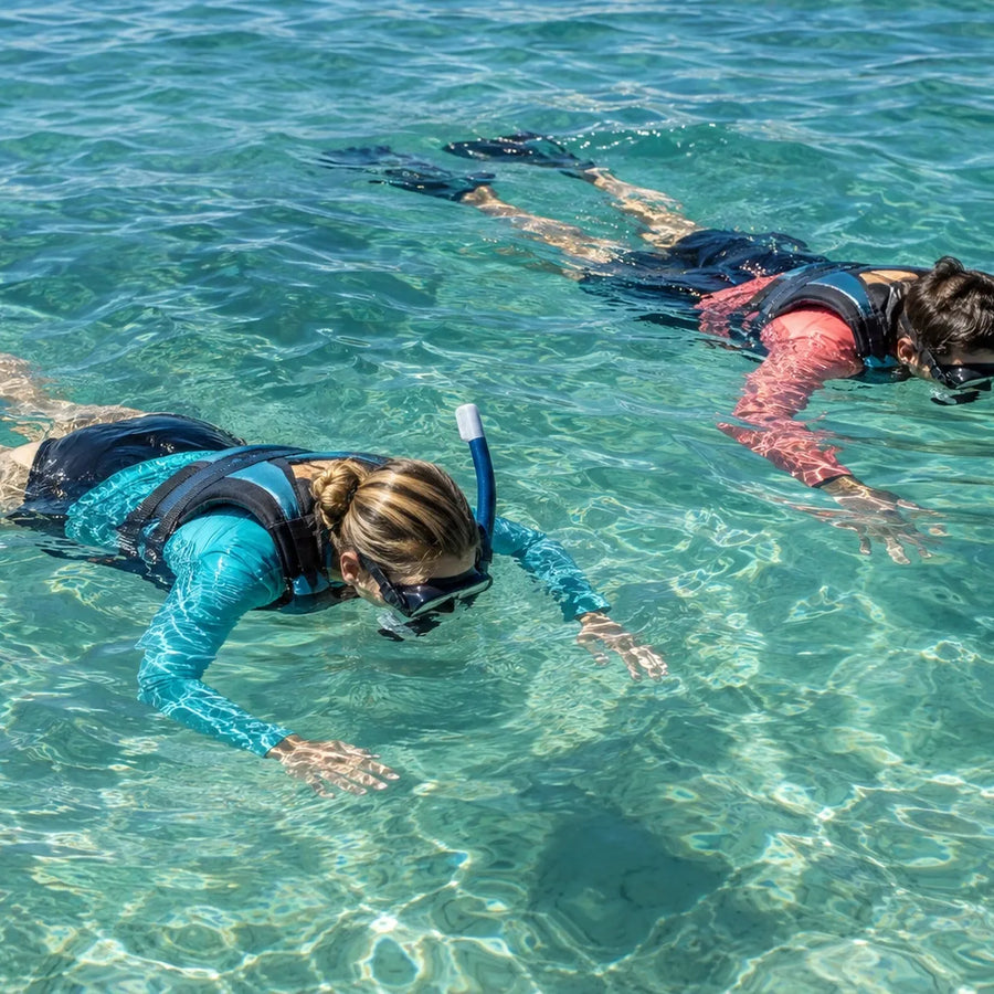Two adults wearing teal and coral swim tops, flotation vests, masks and snorkels float above a sunlit sandy bottom.