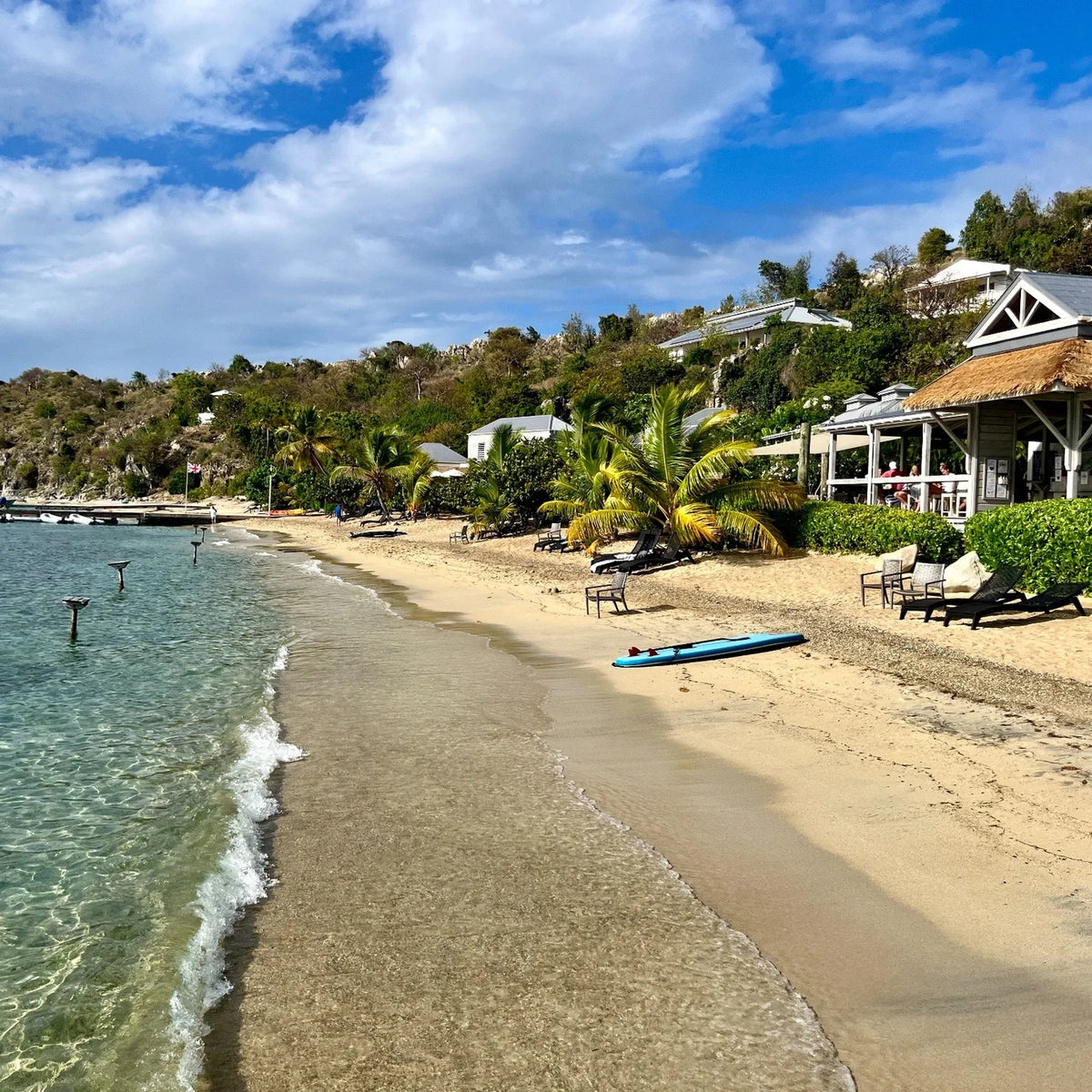 Gentle waves follow a curved sandy beach beside palms, lounge chairs and an open-sided waterfront building, with a blue paddleboard on the sand.