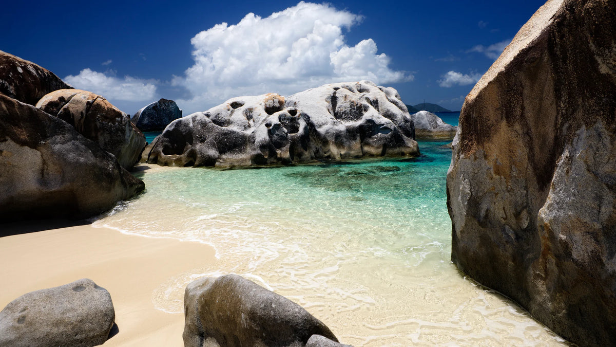 Large rounded granite boulders framing a sandy beach and shallow turquoise water beneath a blue sky.