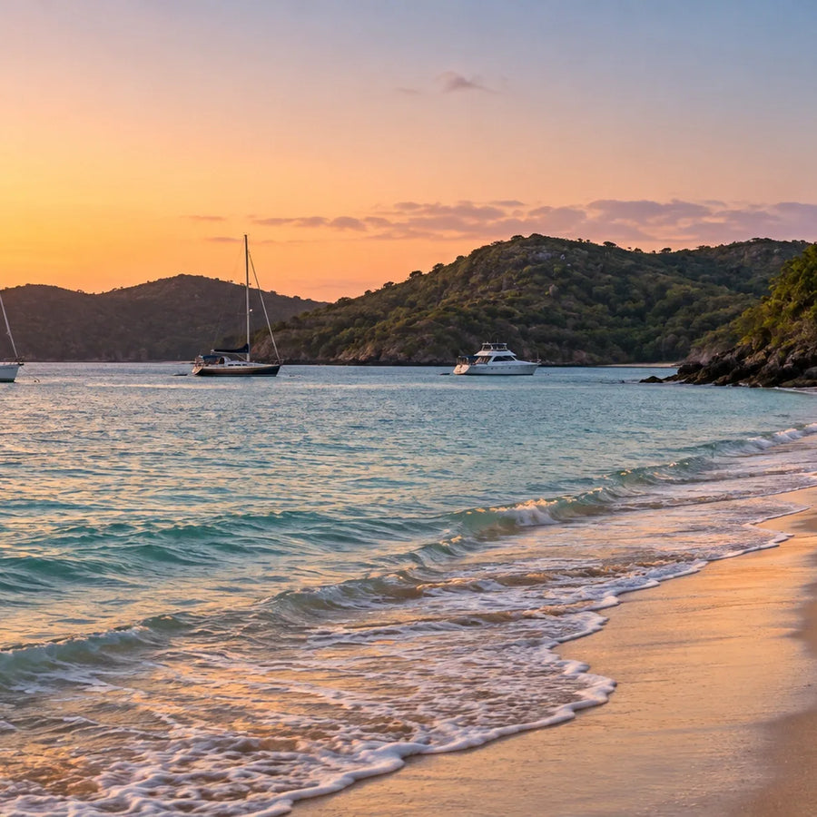 Two adults walk along a sandy beach at dusk, with two sailboats, a power cruiser and wooded hills across the water.