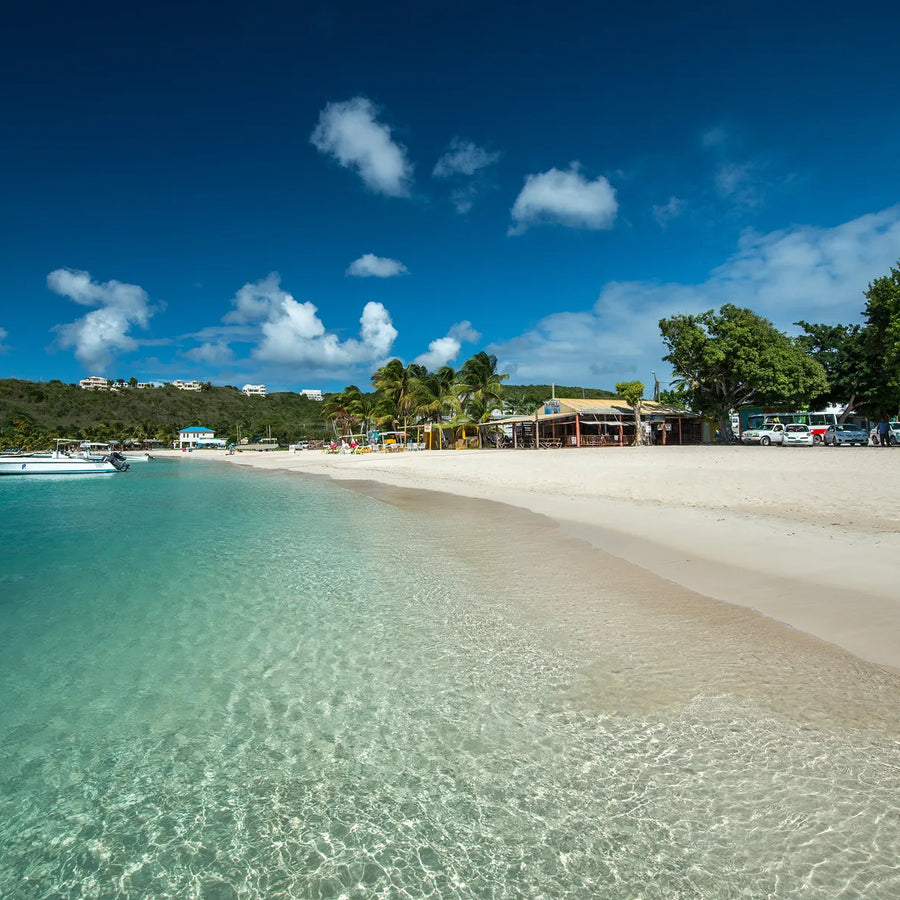 Beach-level view of turquoise water over rippled sand, small boats to the left, and beachfront buildings beneath palms and leafy trees.