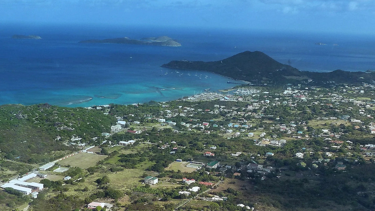Aerial view of a marina with sailboats beside a coastal settlement, green hills, turquoise water and offshore islands.