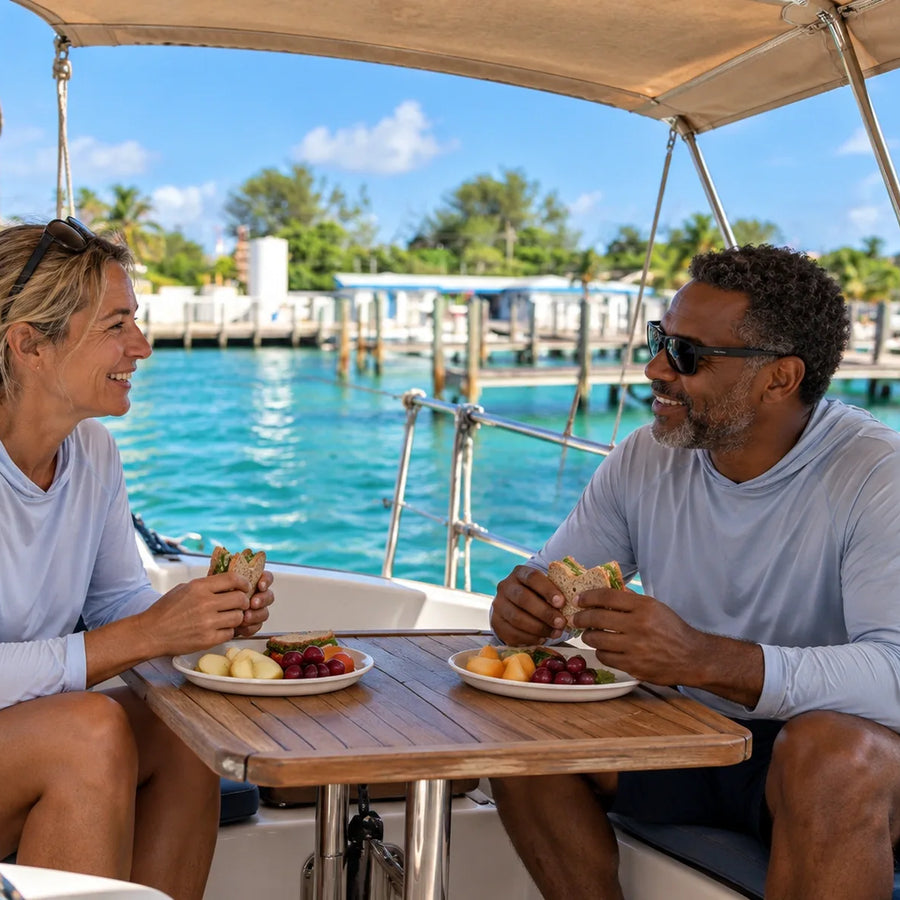 Two boaters share sandwiches and fruit beneath a cockpit shade, with blue-green water and marina docks behind them.