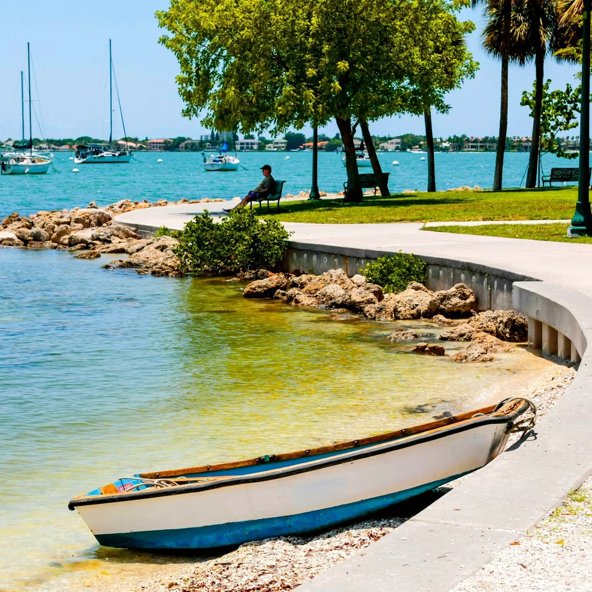 White-and-blue dinghy beside a curved waterfront walkway, with shaded benches and sailboats on turquoise water.