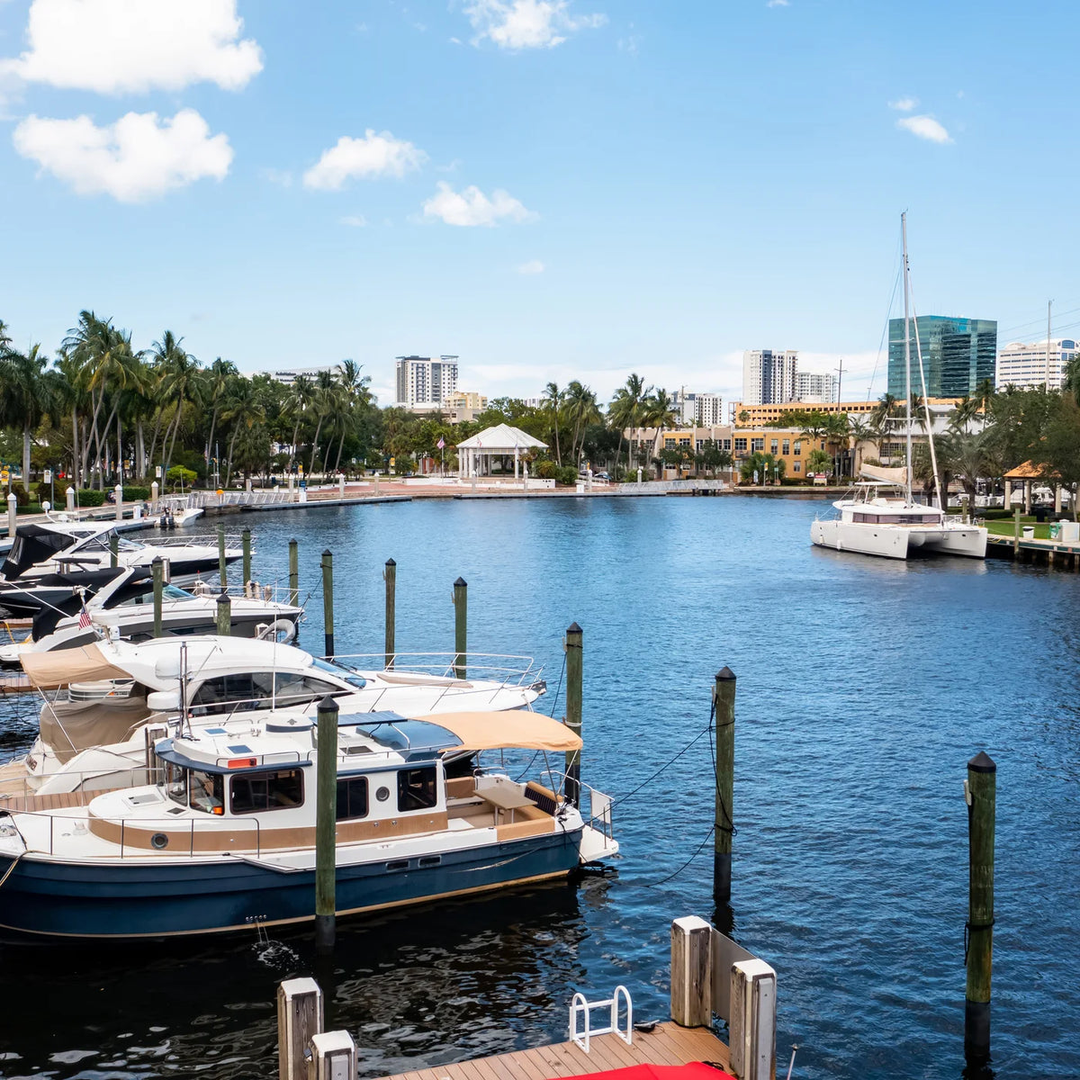 Small cabin boats sit beside docks in the foreground, with a sailing catamaran across the water and palms along the shore.