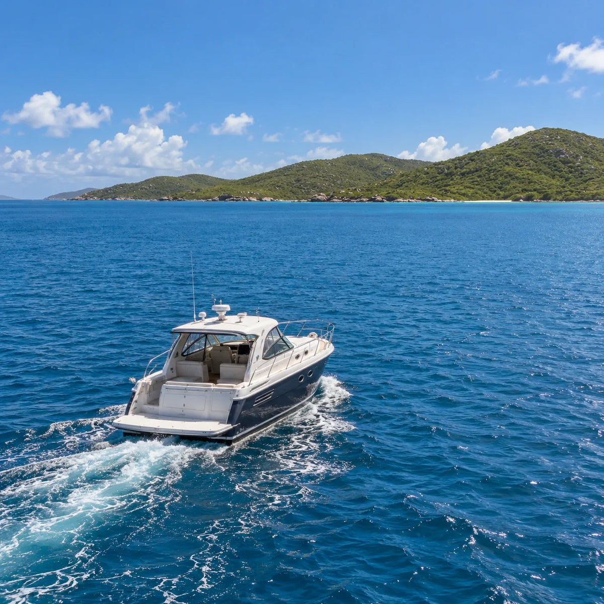 Rear three-quarter view of a power cruiser crossing blue water, with a foamy wake and low coastal hills ahead.