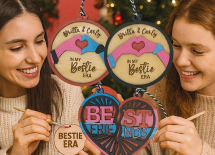 Two smiling young women sit at a holiday crafting table painting wooden best friend ornaments. Above them, colorful text reads “PERSONALIZED YOUR NAMES.” They hold round and heart-shaped wooden ornaments engraved with phrases like “Britta & Carly – In My Bestie Era” and “Best Friends.” The festive background includes a decorated Christmas tree and warm lighting, emphasizing the personalized DIY holiday theme.