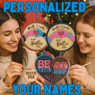 Two smiling young women sit at a holiday crafting table painting wooden best friend ornaments. Above them, colorful text reads “PERSONALIZED YOUR NAMES.” They hold round and heart-shaped wooden ornaments engraved with phrases like “Britta & Carly – In My Bestie Era” and “Best Friends.” The festive background includes a decorated Christmas tree and warm lighting, emphasizing the personalized DIY holiday theme.