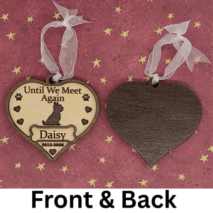 Two heart-shaped wooden dog memorial ornaments shown front and back on a pink background with gold stars. The front ornament is engraved with “Until We Meet Again,” featuring a dog silhouette, paw prints, hearts, the name “Daisy,” and dates “2012–2025.” The back ornament displays the smooth dark wood grain. Both have white ribbons for hanging. Text below reads “Front & Back.”
