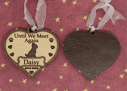 Two heart-shaped wooden dog memorial ornaments shown front and back on a pink background with gold stars. The front ornament is engraved with “Until We Meet Again,” featuring a dog silhouette, paw prints, hearts, the name “Daisy,” and dates “2012–2025.” The back ornament displays the smooth dark wood grain. Both have white ribbons for hanging. Text below reads “Front & Back.”