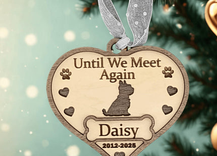 Heart-shaped wooden dog memorial ornament engraved with “Until We Meet Again,” featuring a dog silhouette, paw prints, hearts, and the name “Daisy” with dates “2012–2025.” The ornament hangs from a silver ribbon on a softly lit Christmas tree background. Text below reads “Personalized Dog Memorial Ornament – Custom Wooden Pet Loss Keepsake Gift.”
