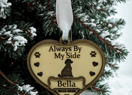Heart-shaped wooden dog memorial ornament engraved with “Always By My Side,” featuring a dog silhouette, paw prints, and hearts. The ornament includes the name “Bella” and dates “2010–2025,” hanging from a white ribbon on a snow-covered evergreen tree branch in a winter setting.