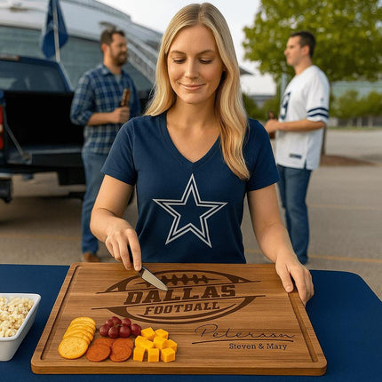 “Blonde woman in a navy Dallas football shirt prepares snacks on a personalized Dallas Football acacia cutting board engraved with the name ‘Peterson – Steven & Mary’ at a tailgating party. The board holds crackers, pepperoni, grapes, and cheese cubes, with fans, a truck, and the stadium visible in the background.”