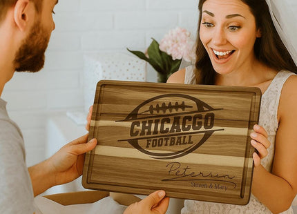 “Smiling bride receiving a personalized Chicago football cutting board as a wedding gift from her partner. The engraved acacia wood board features a football logo and the family name ‘Peterson – Steven & Mary.’ The couple sits indoors surrounded by gift wrap, highlighting the board as a thoughtful and unique wedding present.”