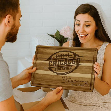 “Smiling bride receiving a personalized Chicago football cutting board as a wedding gift from her partner. The engraved acacia wood board features a football logo and the family name ‘Peterson – Steven & Mary.’ The couple sits indoors surrounded by gift wrap, highlighting the board as a thoughtful and unique wedding present.”